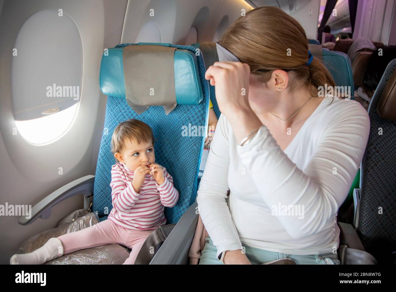 Bequemer Flug mit Kind. Niedliches kleines Kleinkind und Mama sitzen auf dem Flugzeug in Stühlen und schauen sich gegenseitig an. Mama hebt eine Schlafmaske Stockfoto