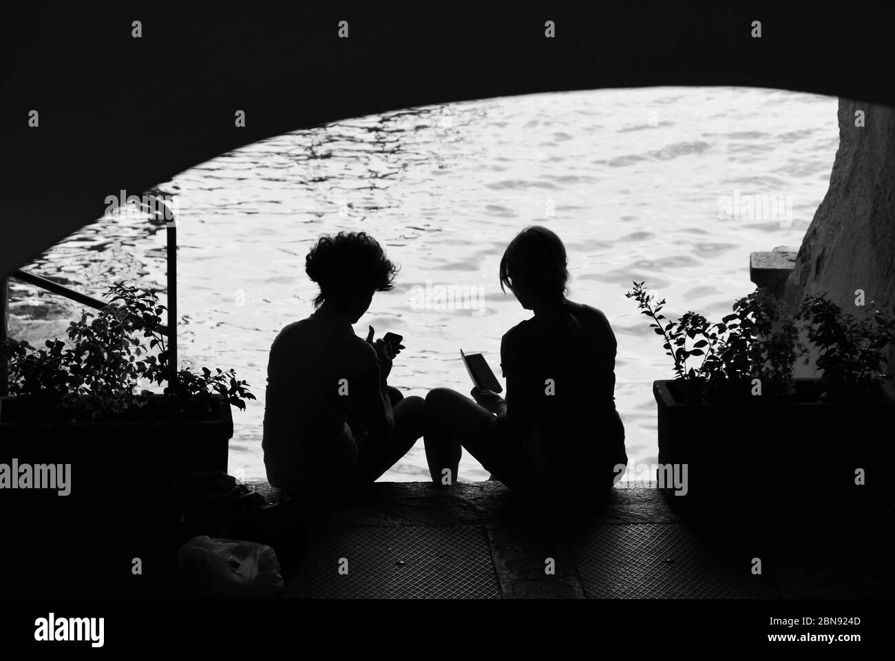 VARENNA, COMER SEE, ITALIEN - JUNI 2019: Silhouette zweier junger Frauen, die am Ufer des Wassers unter einem Gebäude in Varenna am Gardasee sitzen Stockfoto