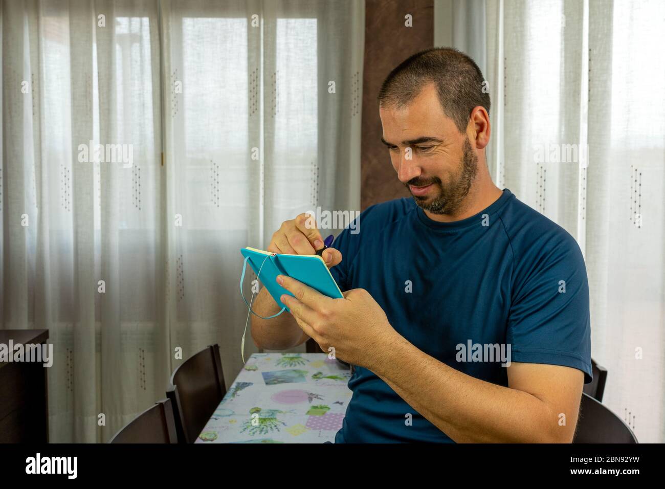 Lächelnder Mann mit Bart und kurzen Haaren zeigt Notizen auf seinen kleinen blauen Notizblock im Wohnzimmer zu Hause Stockfoto