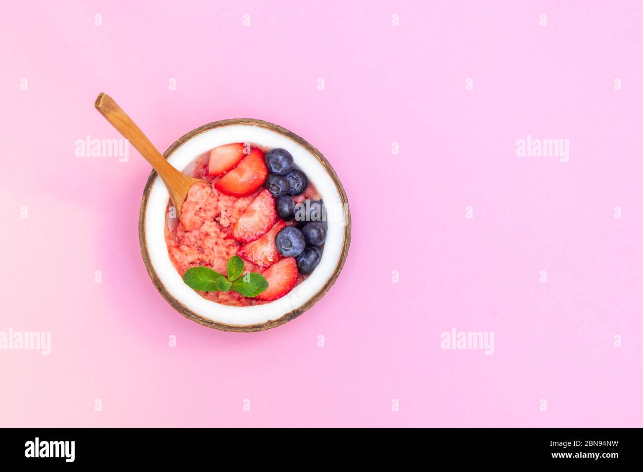 Gesundes vegetarisches Dessert. Draufsicht der Kokosnussschale mit Beerensoothie auf weichem rosa Hintergrund, Kopierraum Stockfoto