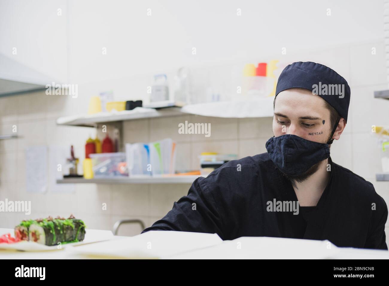 Porträt eines Sushi-Küchenchefs mit Gesichtsmaske in der kommerziellen Küche. Essen zum Mitnehmen, Image des lokalen Catering-Unternehmens Stockfoto