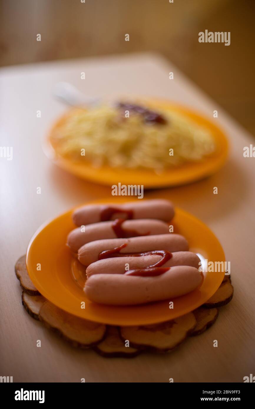 Fünf Würste auf einem orangefarbenen Teller, auf einem Hintergrund ein Gericht mit Spaghetti Stockfoto
