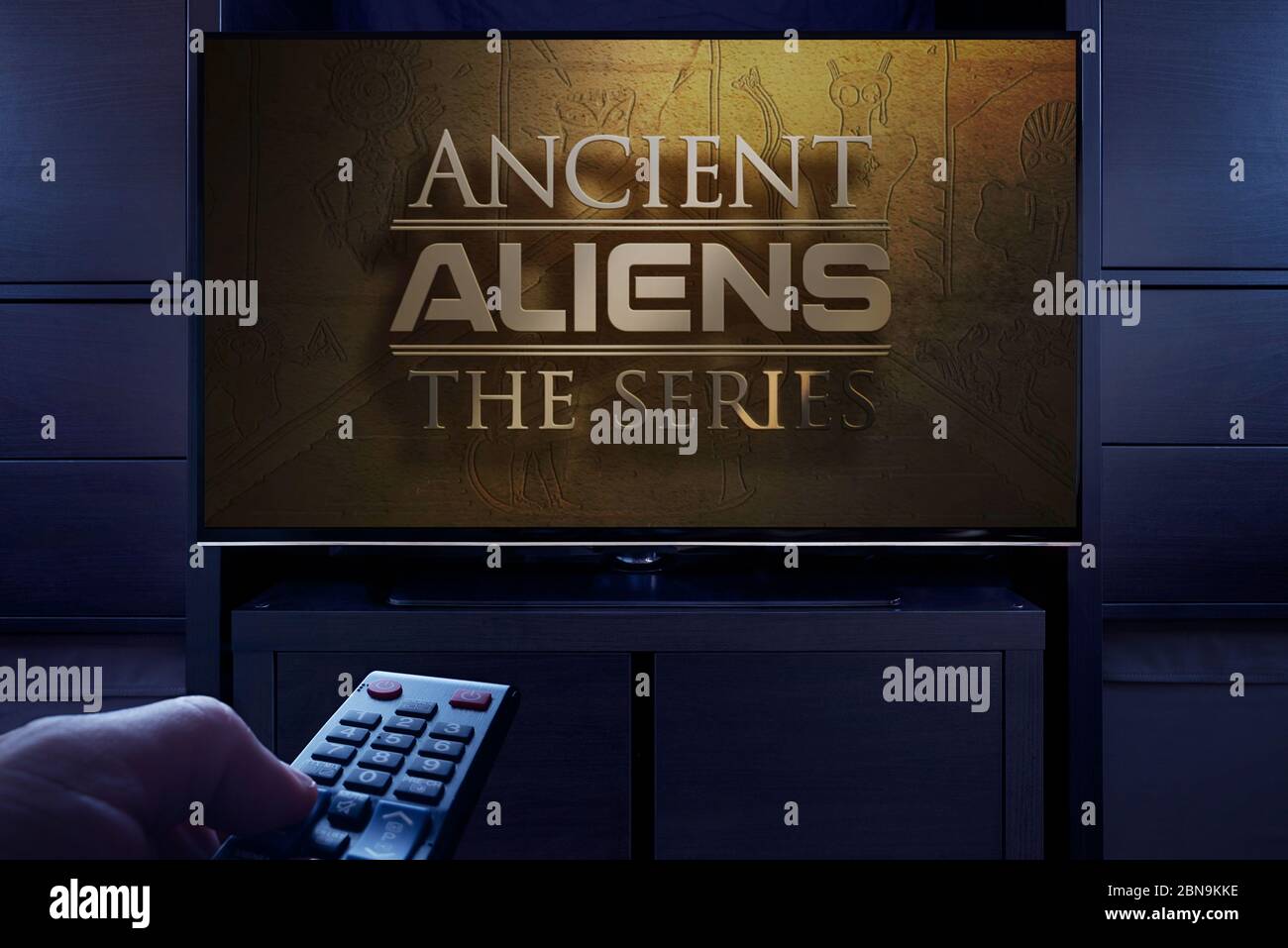Ein Mann zeigt mit einer Fernbedienung auf den Fernseher, auf dem der Titelbildschirm von Ancient Aliens angezeigt wird (nur redaktionelle Verwendung). Stockfoto