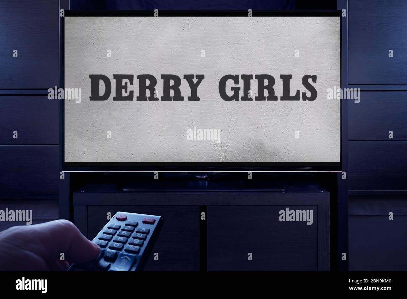 Ein Mann zeigt eine Fernbedienung auf den Fernseher, der den Hauptbildschirm von Derry Girls anzeigt (nur redaktionelle Verwendung). Stockfoto