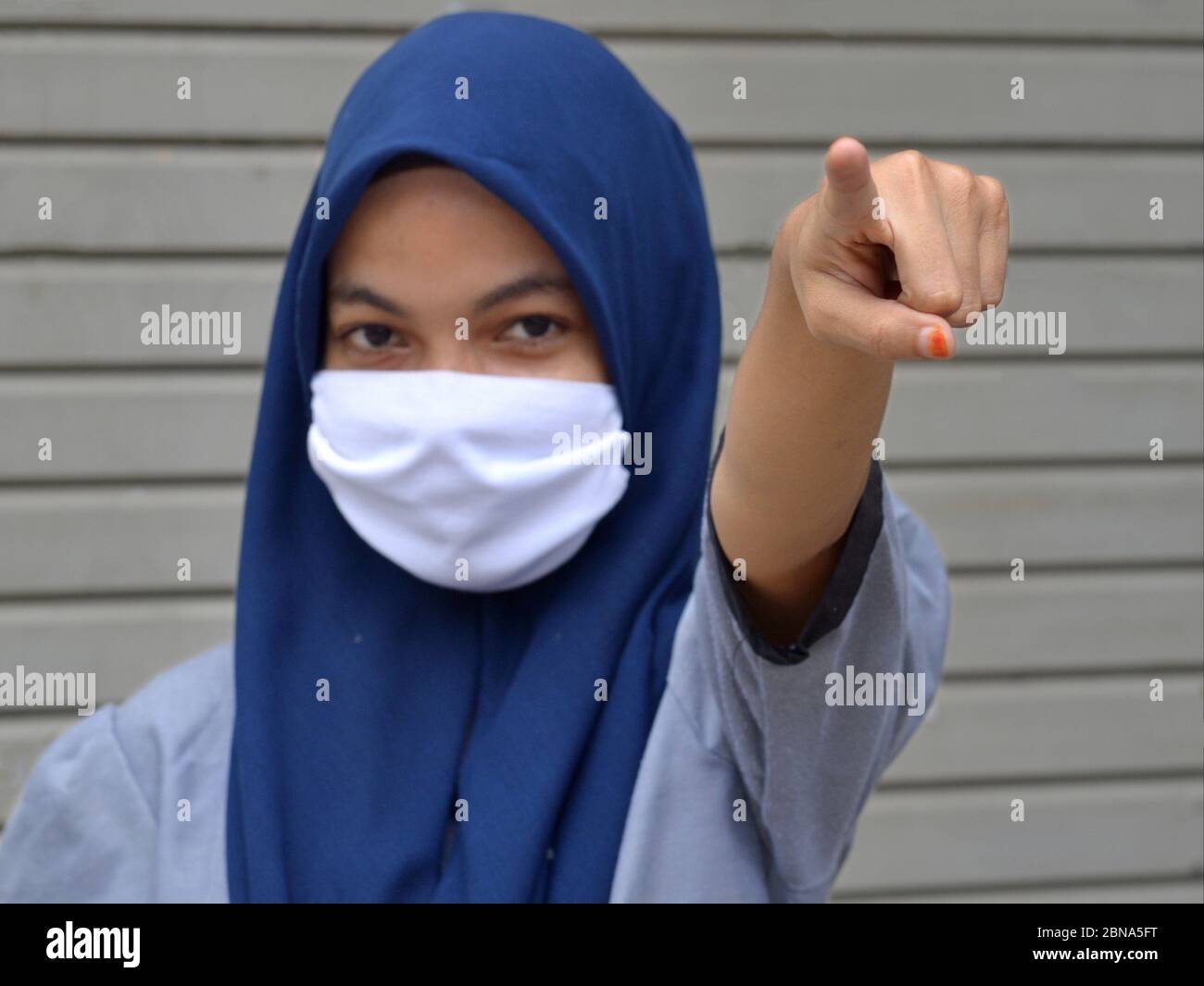 Muslimische malaiische Mädchen mit Hijab und Mund-deckenden Schleierpunkten während der Corona-Pandemie mit ihrem Zeigefinger zum Betrachter: Halten Sie Ihre soziale Distanz! Stockfoto