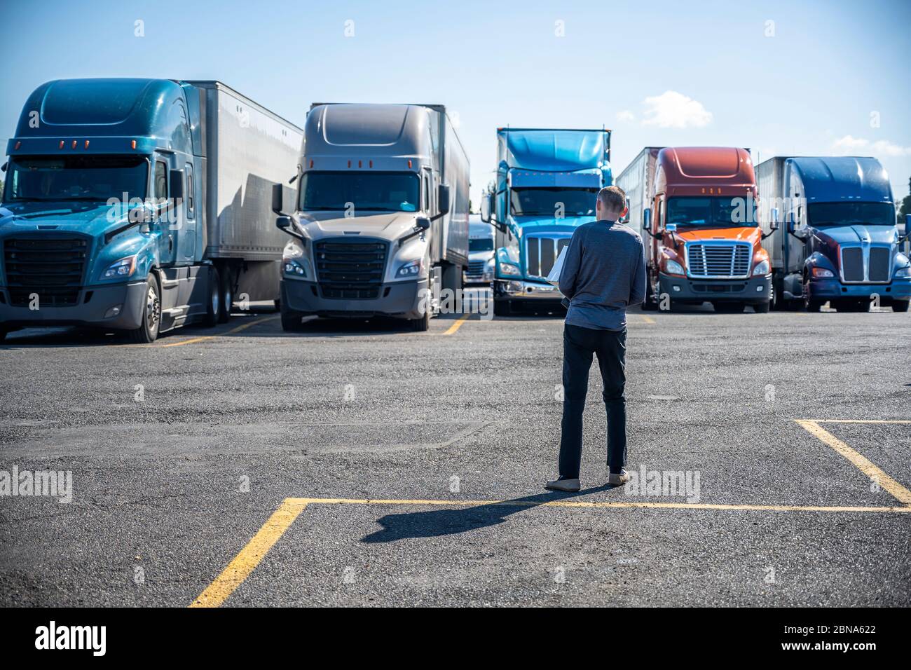 Slim Truck Fahrer trägt eine Box mit Käufen in seinen Händen und geht zu seinem großen Rig semi Ruck auf dem LKW-Stopp-Parkplatz in Reihe wi geparkt Stockfoto