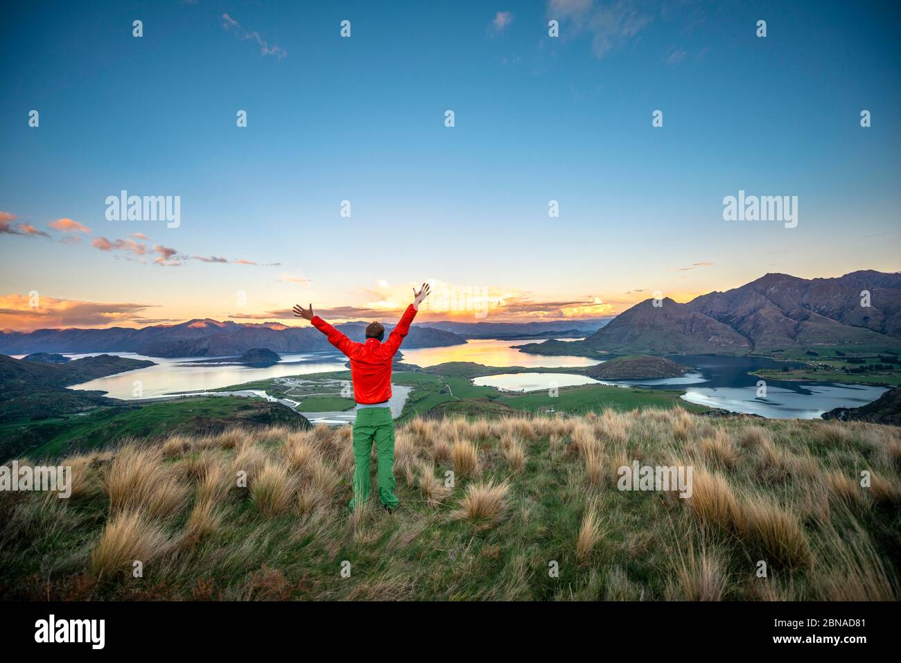 Wanderer streckt seine Arme in der Luft, Panoramablick auf Wanaka Lake und Berge bei Sonnenuntergang, Rocky Peak, Glendhu Bay, Otago, South Island Stockfoto