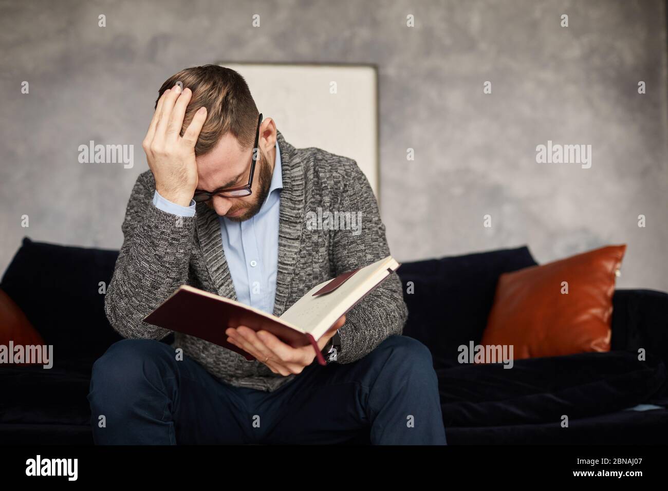 Junger Geschäftsmann hält seinen Kopf, während er auf dem Sofa sitzt und seine Pläne und Aufgaben im Notizblock im Büro liest Stockfoto