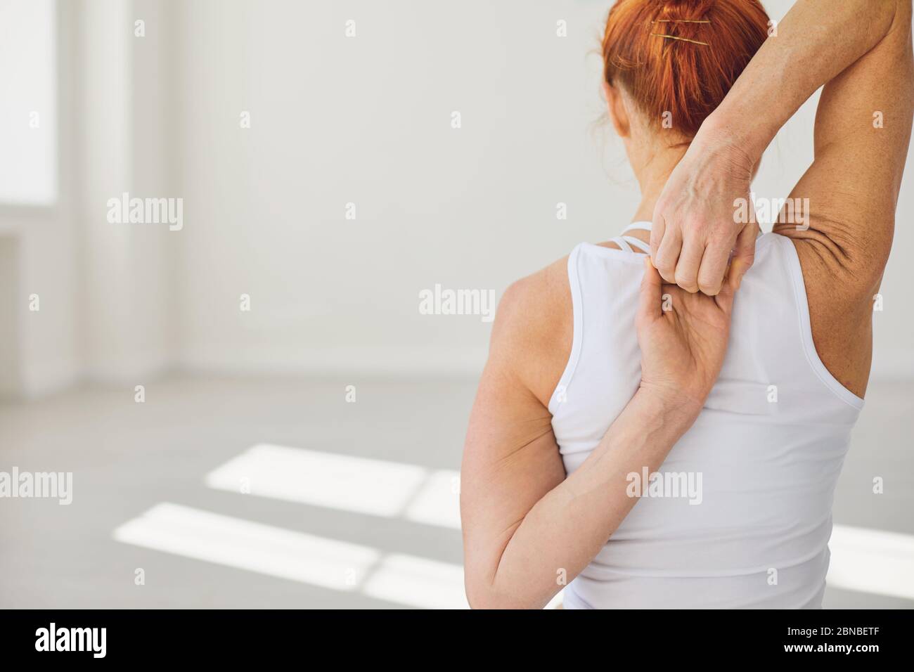 Rückansicht der jungen fit Frau in weißer Sportbekleidung sitzt in Gomukhasana Pose mit den Händen hinter dem Rücken während Yoga in Licht Studio üben Stockfoto
