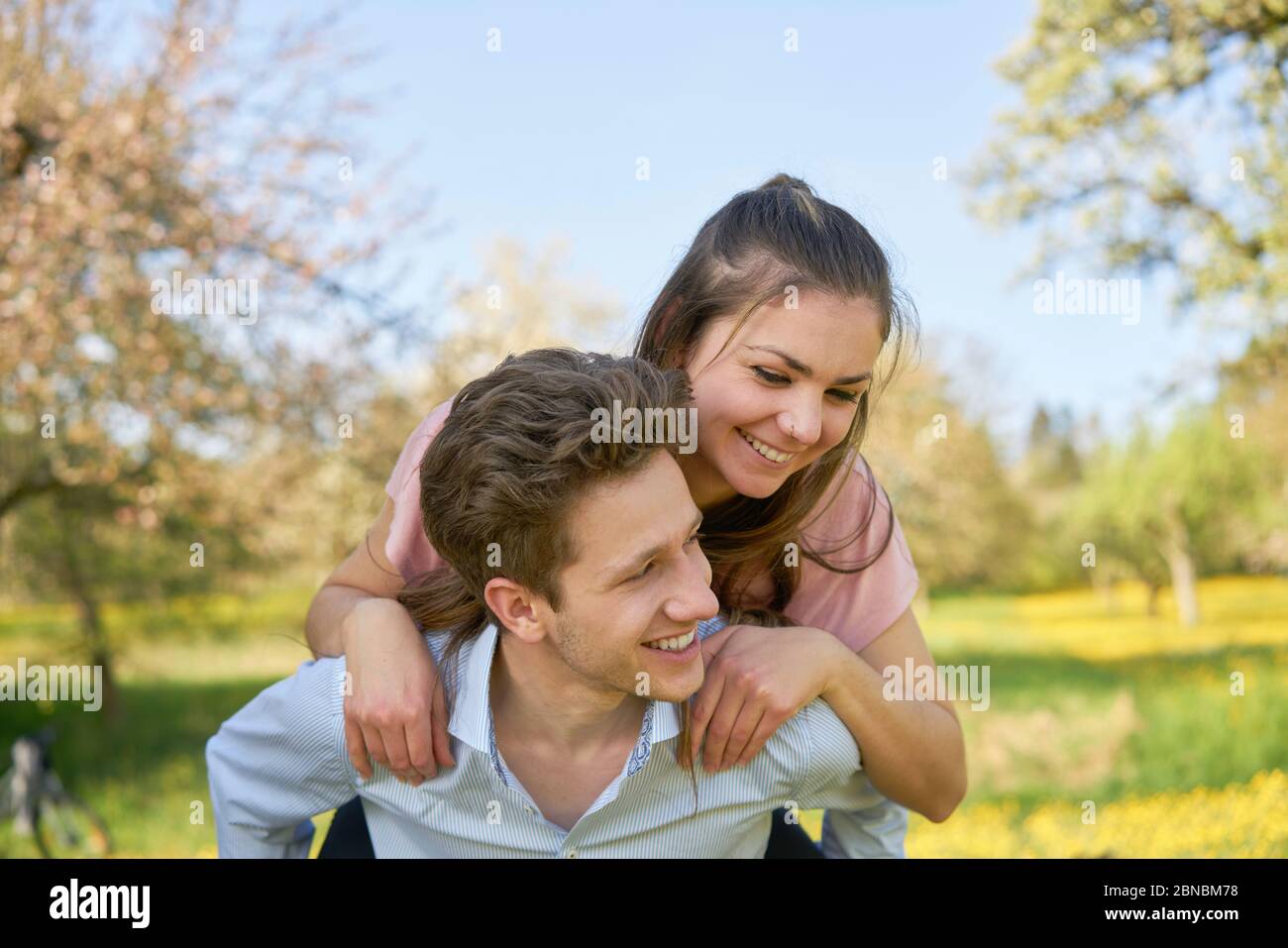 Junges Paar in der Natur, ein Freund, der seine Freundin auf dem Rücken auf einer Wiese mit gelben Blumen und Bäumen trägt, im Hintergrund ein eisiger Stockfoto