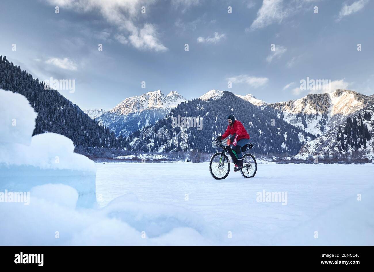 Der Mann in roter Jacke fährt mit seinem Fahrrad am gefrorenen See in verschneiten Bergen im Hintergrund Stockfoto
