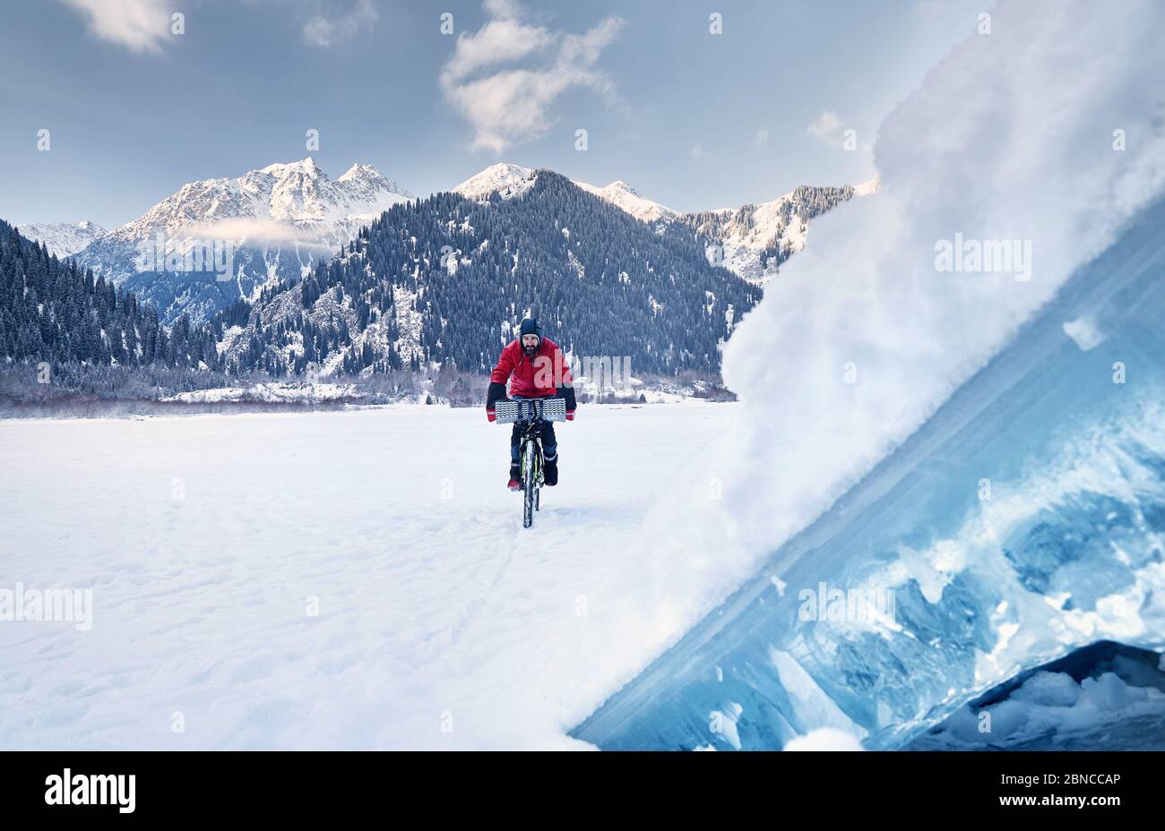 Der Mann in roter Jacke fährt sein Fahrrad am gefrorenen See mit blauem Eis im Vordergrund in verschneiten Bergen im Hintergrund Stockfoto
