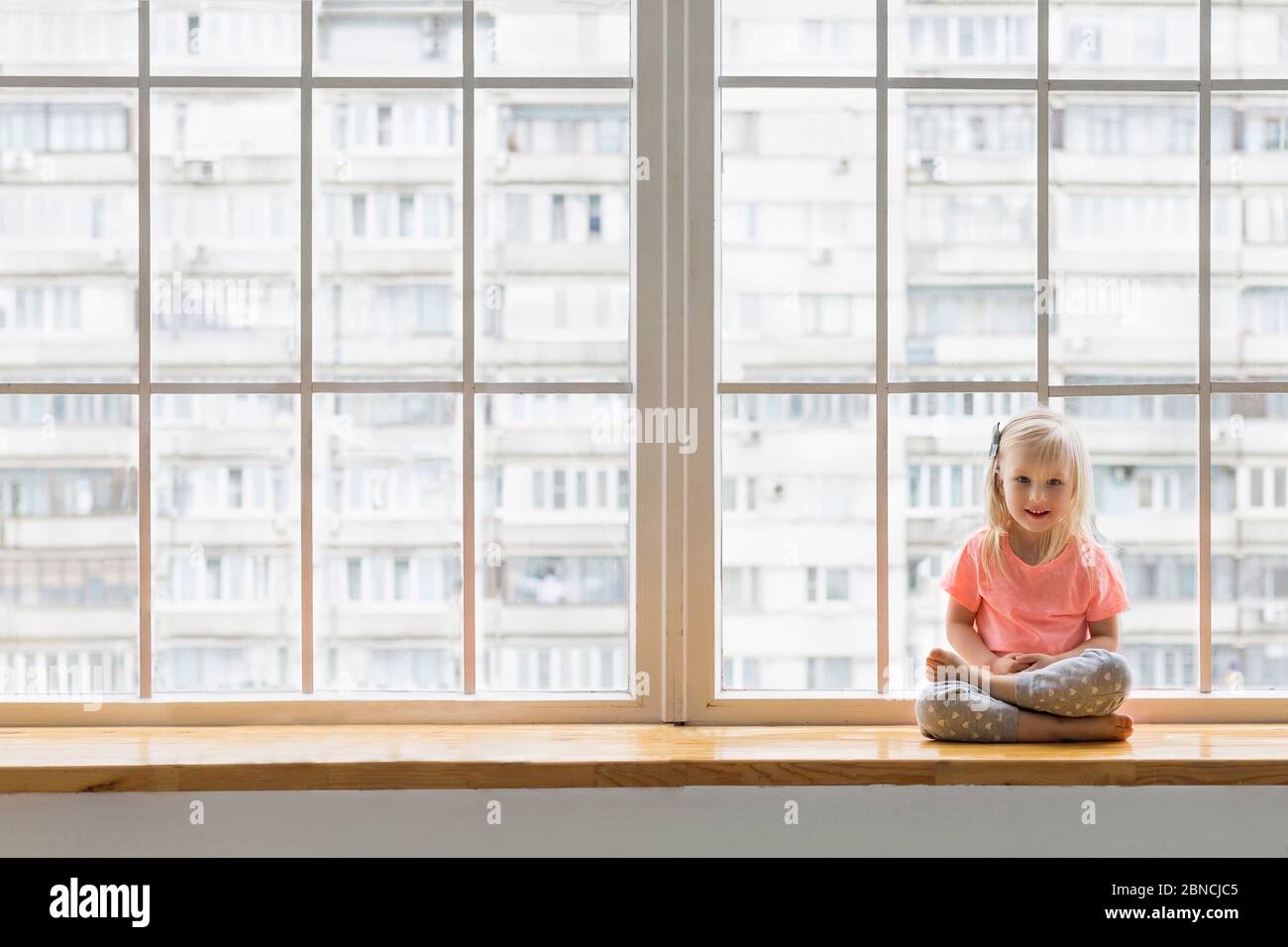 Nettes 3-4 Jahre altes Mädchen, das in Lotushaltung mit gekreuzten Beinen auf der Fensterbank sitzt Stockfoto