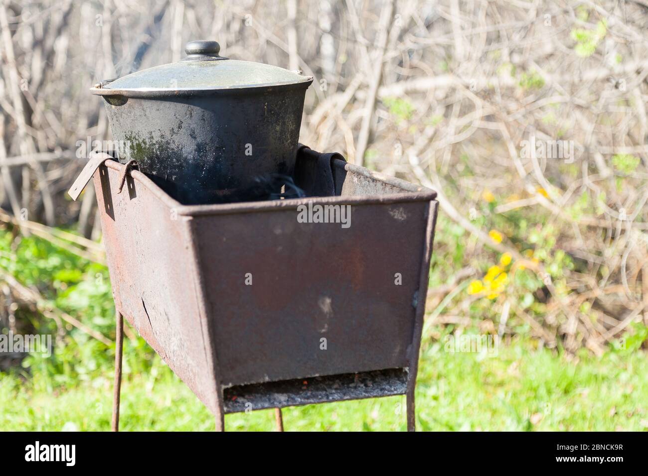 Schwarzer geräucherter gusseiserner Kessel mit Deckel auf Kohlen in einem Metallfass vor grünem Gras. Kochen Pilaf mit handlichen Mitteln auf einem offenen Stockfoto