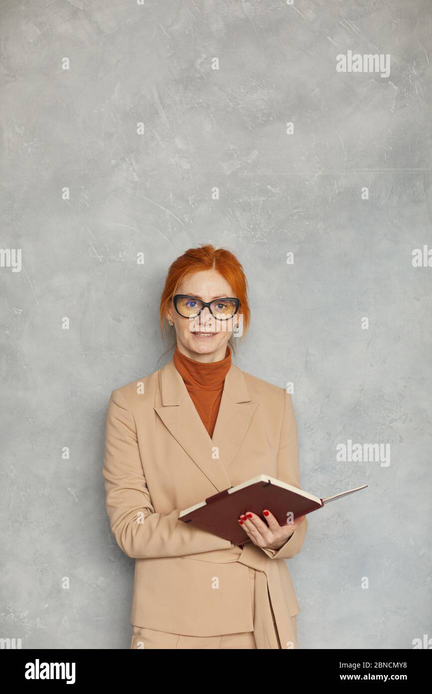 Porträt einer rothaarigen reifen Geschäftsfrau in Brillen, die ihr Notizblock hält und die Kamera gegen die graue Wand lächelt Stockfoto