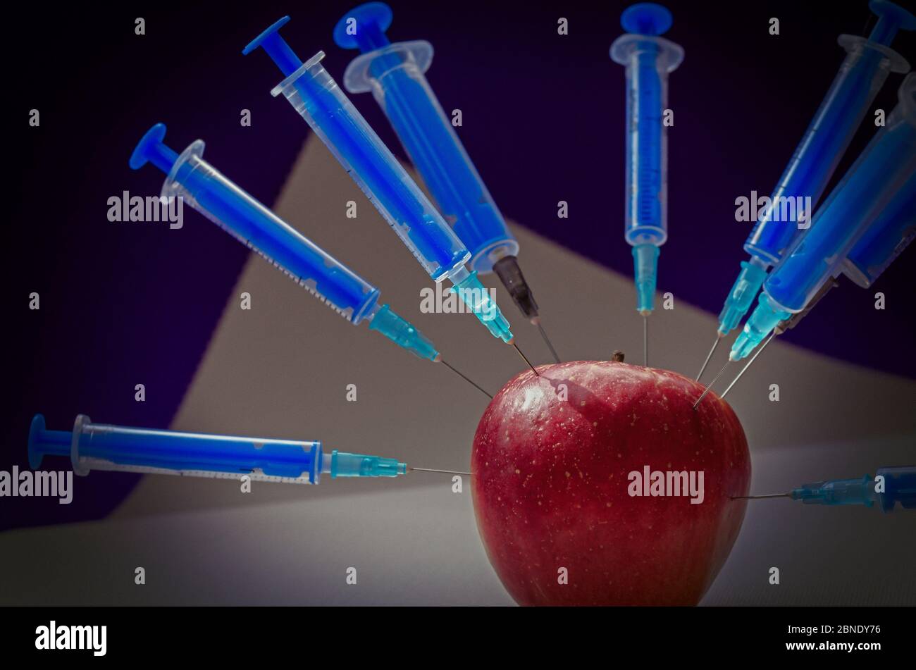 Apfel mit Spritzen besetzt. Reifer roter apfel von allen Seiten von Spritzen durchbohrt. Gesundheitswesen oder Kosmetologie. Mehrdeutiger Hintergrund. Seitenansicht. Selektiv Stockfoto