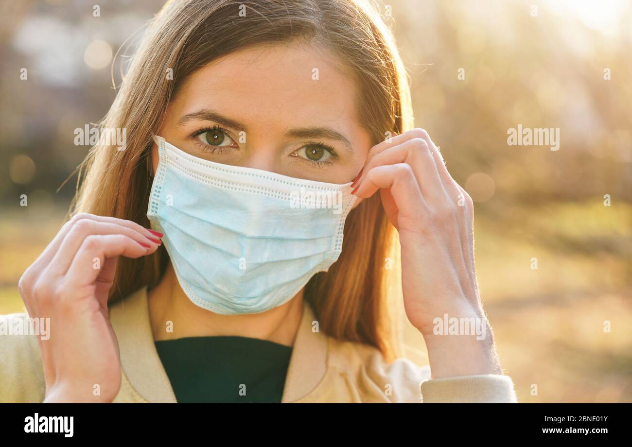 Junge Frau trägt blaue Einweg-Virus Mund Nasenmaske, berühren sie mit den Händen, schöne sonnige Bokeh im Hintergrund, Nahaufnahme Gesicht Porträt Stockfoto