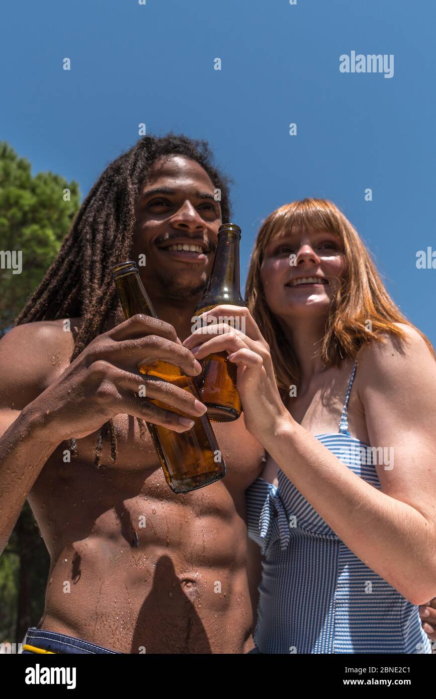 Vertikales Foto einer jungen Frau neben einem afrikanischen Mann mit Dreadlocks, die mit einer Flasche Bier in einem Außenpool toasten Stockfoto
