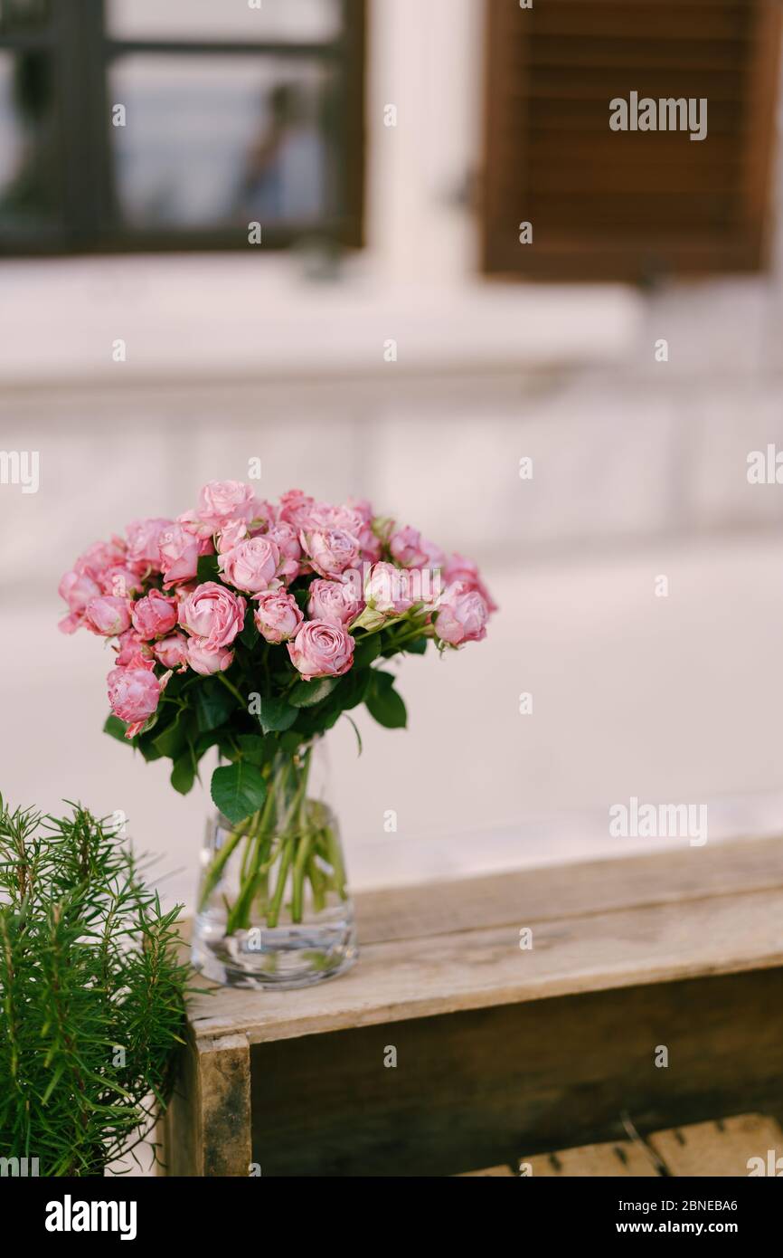 Empfang am Tisch für Hochzeitsessen. Ein Strauß rosa Rosen in einer gläsernen transparenten Vase mit Wasser, steht auf einer Holzkiste vor dem Hintergrund eines Stockfoto
