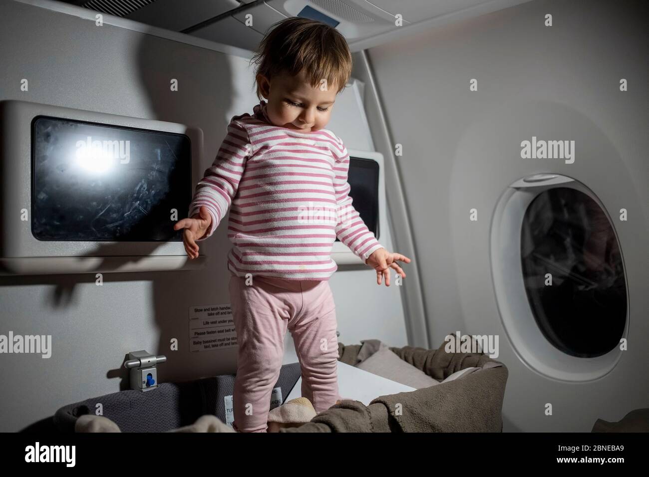 Verletzung der Sicherheitsregeln im Flugzeug. Gefahr. Niedliches kleines Kleinkind steht während des Fluges in Babybettchen Stockfoto