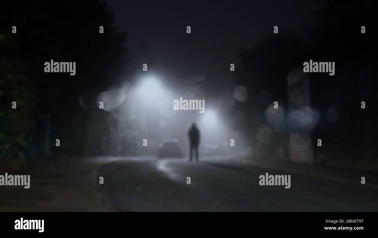 Eine geheimnisvolle Figur, die in einer nebligen Winternacht mitten auf einer Straße mit Straßenlaternen steht. Mit einem unscharfen, Bokeh, unscharf bearbeitenden Schnitt Stockfoto