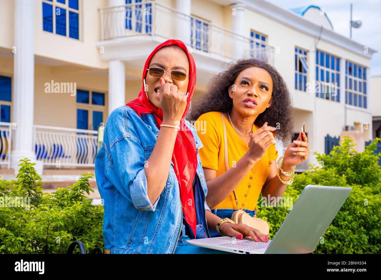 Zwei afrikanische Frauen sitzen draußen in einem Park und kaufen zusammen mit einem Laptop ein, sie sehen erschrocken aus Stockfoto