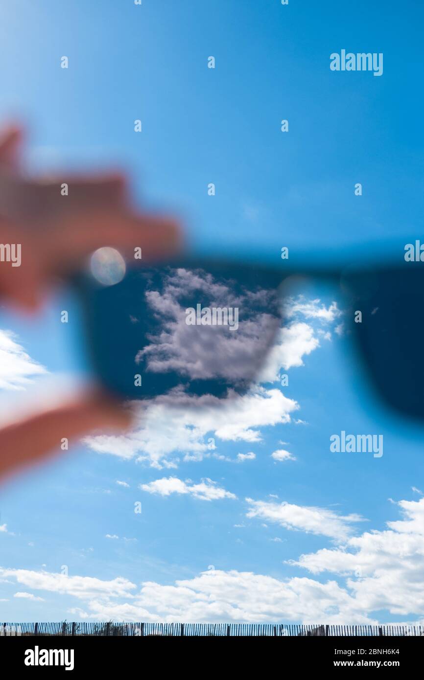 Nahaufnahme der Hand, die die Sonnenbrille zum sonnigen Himmel hält, mit Wolken im Fokus auf der linken Linse Stockfoto