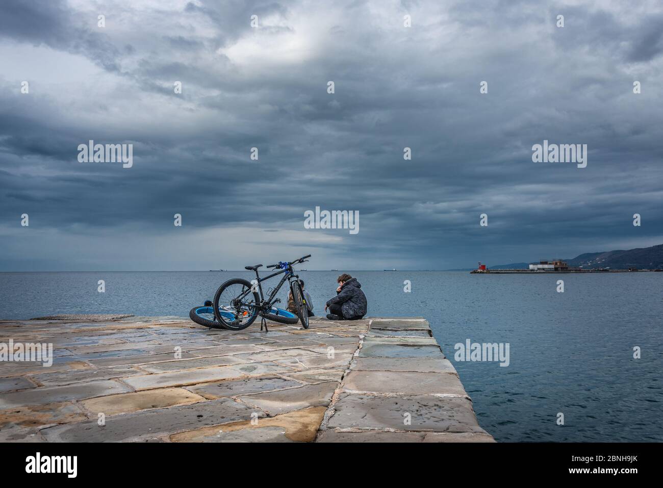 Jugendliche mit Fahrrädern im Molo Audace mit Blick auf die Bucht von Triest mit dramatischem Winterhimmel Stockfoto