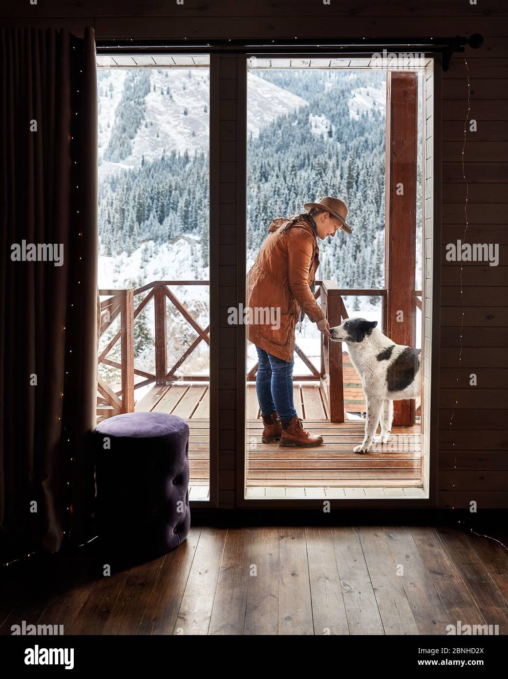 Frau in Hut spielt mit ihrem Hund im Winter im Holzhaus im Bergresort Stockfoto