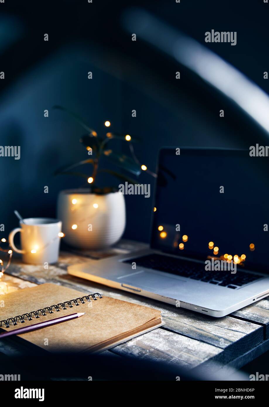 Gemütlicher Arbeitsplatz mit Laptop, Note mit Bleistift und Tasse Kaffee, Pflanzen und gelb glühenden garlandat Stein grauen Wand Hintergrund. Arbeiten von zu Hause Konz Stockfoto