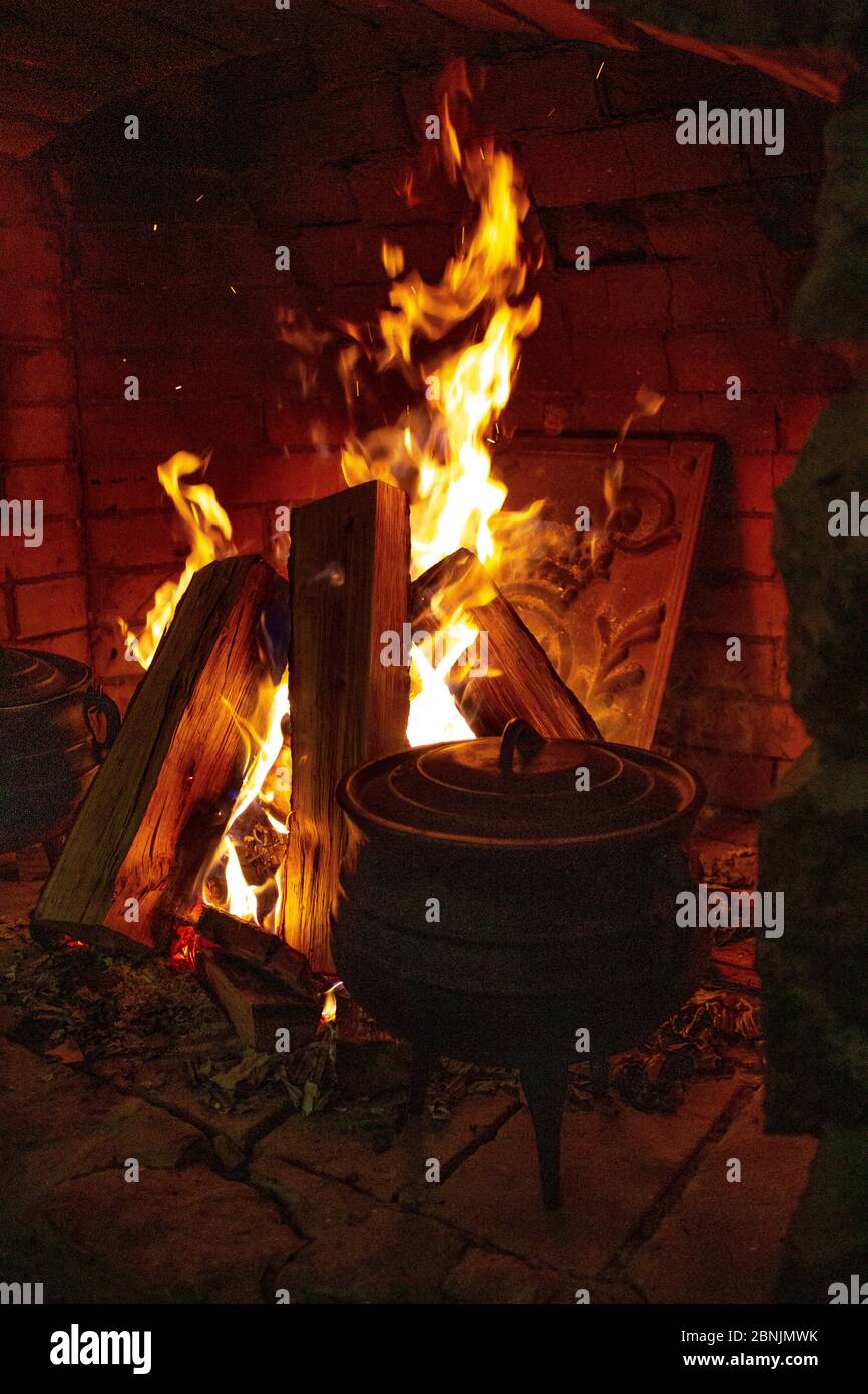 Offenes Feuer im Kamin. Ein Herd, in dem das Essen gekocht wird. Gusseiserne Kessel zum Kochen auf dem Scheiterhaufen. Brennholz im Kamin. Gemütliches Zuhause Stockfoto