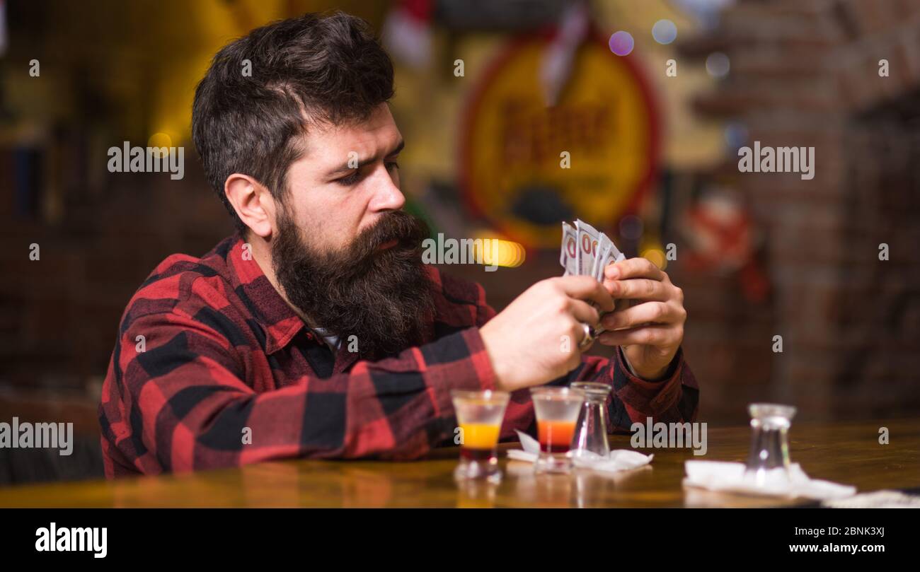 Get Drunk Concept. Mann mit betrunkenem Gesicht sitzen alleine an der Theke. Hipster hält Geld, zählen Bargeld, um mehr Alkohol zu kaufen. Guy verbringen Freizeit in Bar trinken Cocktail, defokussed Hintergrund. Stockfoto