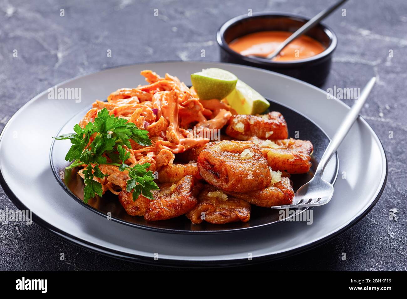 Karibische Küche: Gebratene Kochbananen mit geschreddertem Huhn mit Büffelsauce, serviert mit Limette und Petersilie auf einem schwarzen Teller mit Gabel, Stockfoto