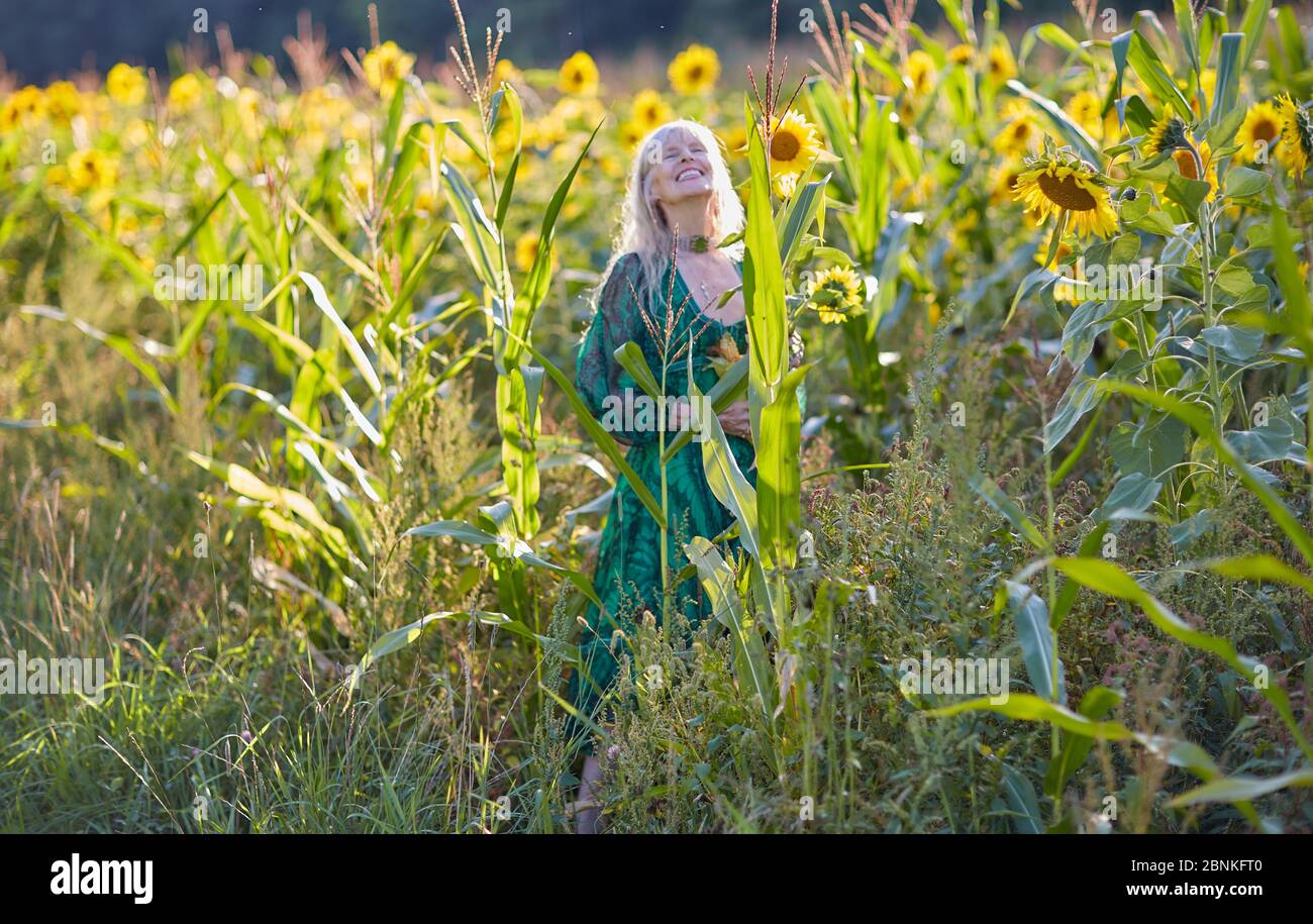 Frau mittleren Alters, die den Himmel betrachtet, lachend, grünes Kleid, Sonnenblumenfeld, Mais auf der Kohle Stockfoto