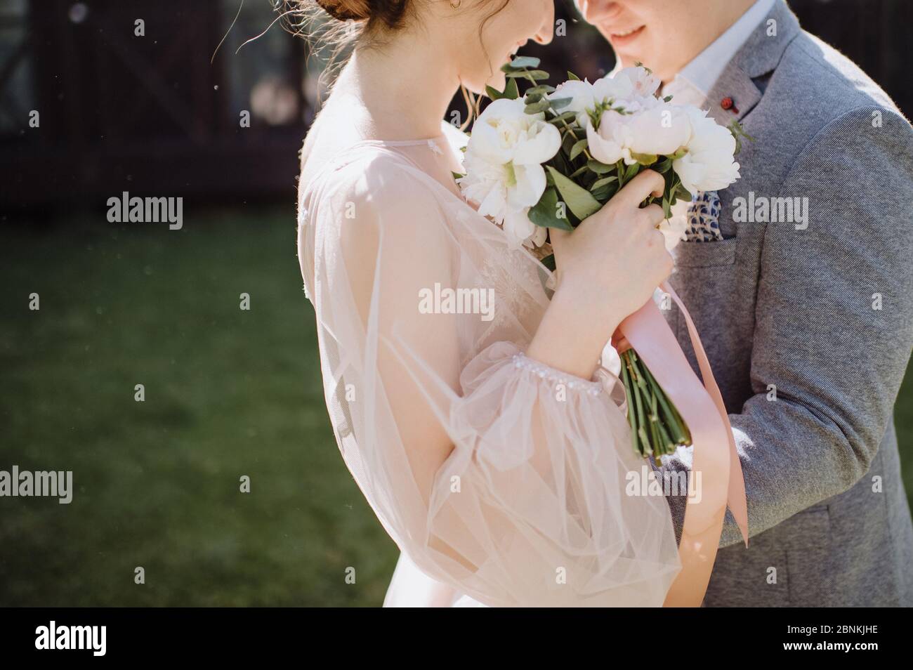 Braut und Bräutigam umarmen sich, stehen einander gegenüber. Die Braut in einem rosa Kleid hält in ihren Händen einen Strauß aus weißen und rosa Pfingstrosen. Der Strauß ist Krawatte Stockfoto