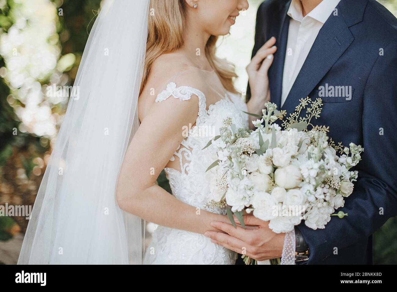 Nahaufnahme des Straußes einer Braut aus weißen Blumen. Die Braut in einem weißen Spitzenkleid hält einen Strauß in den Händen. Die Braut und Bräutigam umarmen, stehen mit Blick auf EA Stockfoto