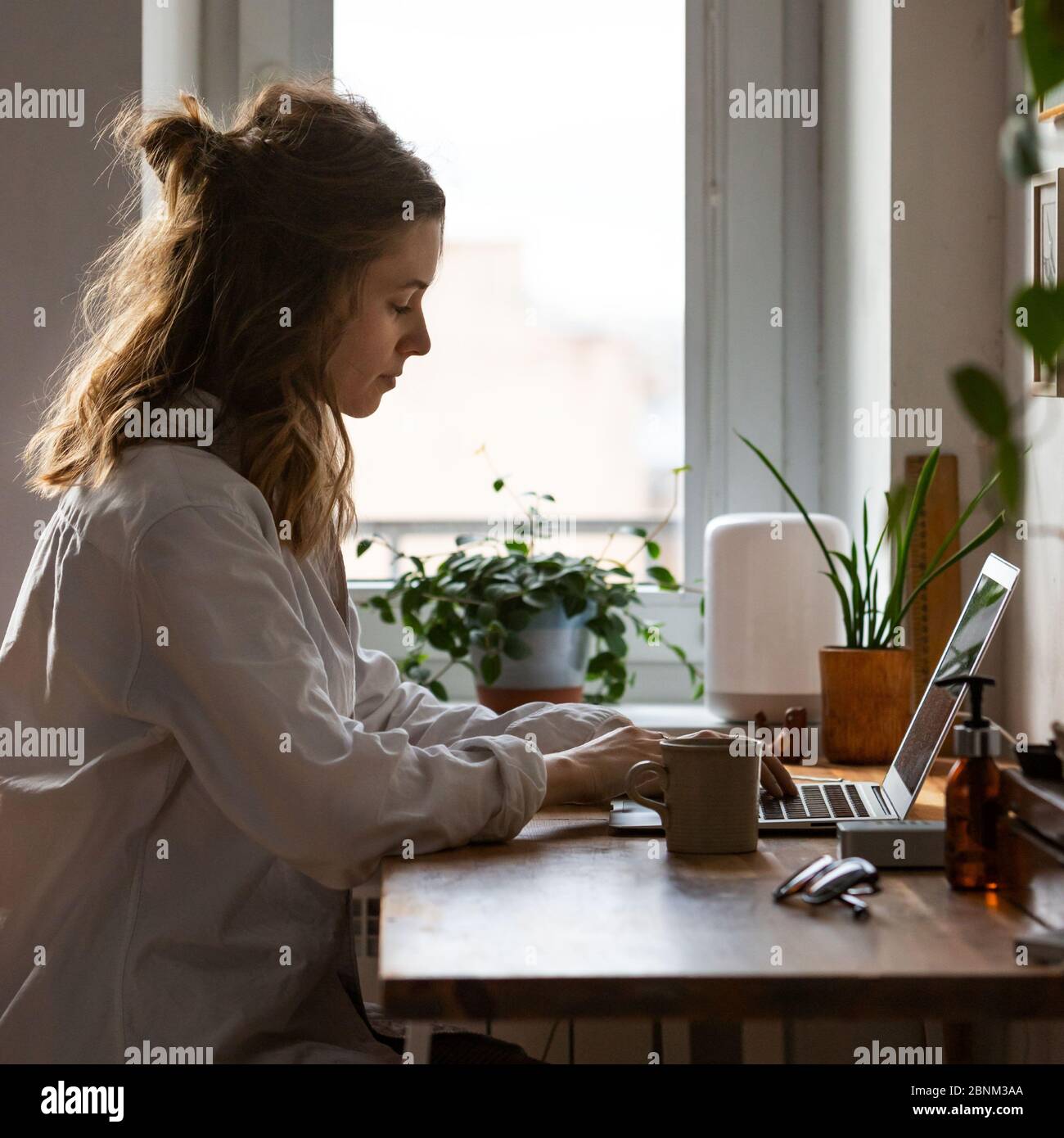 Junge Frau freiberufliche/Designer arbeitet am Computer aus dem Home Office während der Selbstisolierung durch Coronavirus. Gemütlicher Arbeitsplatz umgeben von Pflanzen. Re Stockfoto