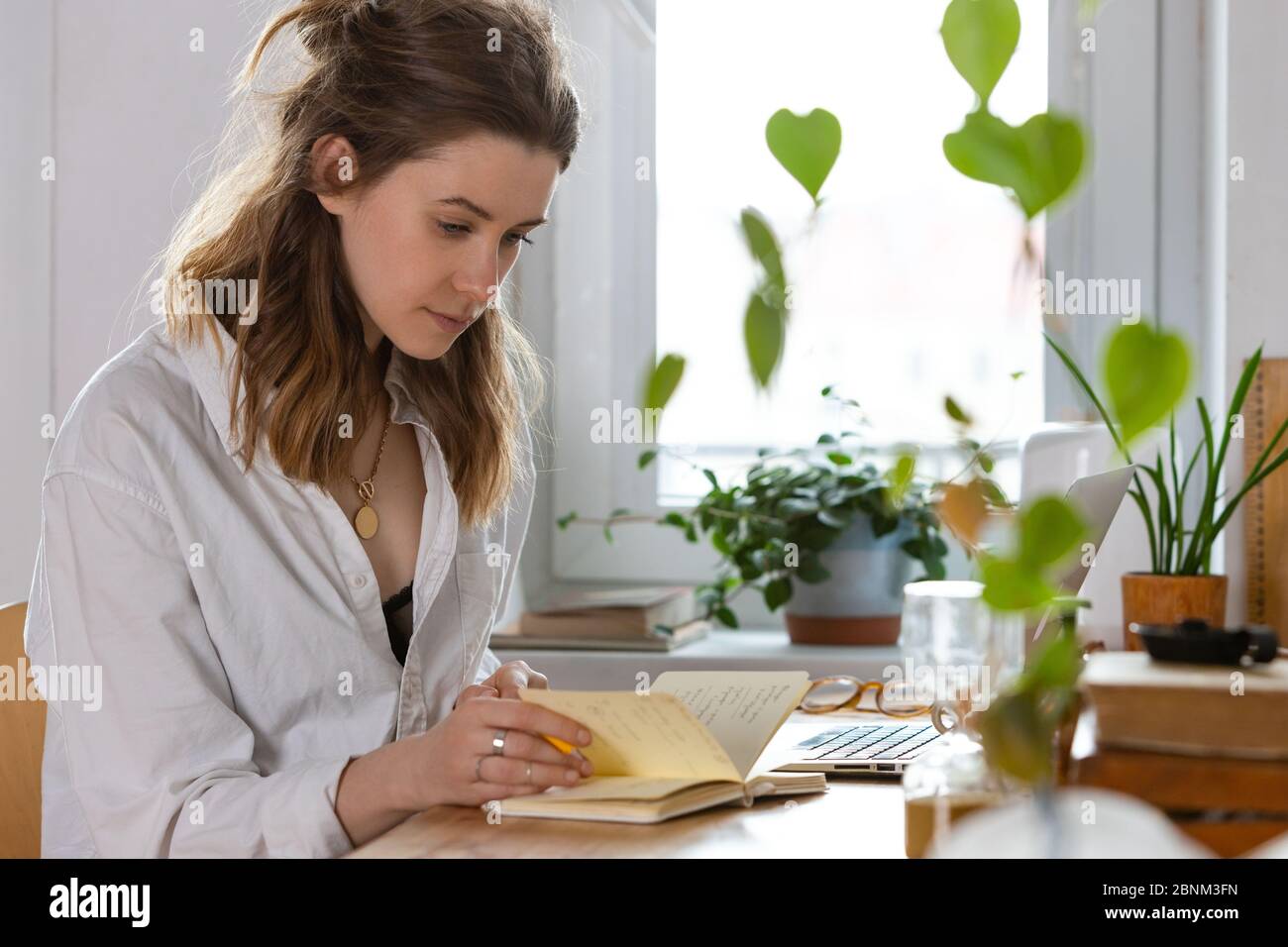 Junge Freiberuflerin/Designerin macht Notizen in einem Notizbuch, die während der Selbstisolierung durch Coronavirus zu Hause arbeitet. Gemütlicher Arbeitsplatz umgeben von Plan Stockfoto