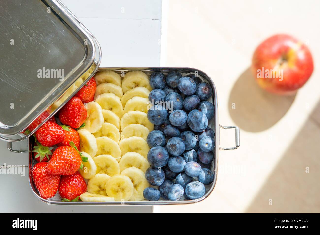 Heidelbeeren, Bananen und Erdbeeren machen einen Snack in einem großen Edelstahl-Lebensmittelbehälter. Plastikbox für Mittagessen auf dem Tisch isoliert. Roter Apfel auf dem Hintergrund. Keine Verschwendung, umweltfreundlich. Stockfoto