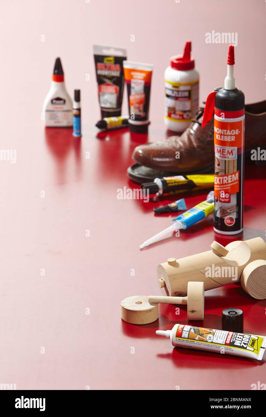 Stillleben Studiofotografie zum Thema Kleben. Verschiedene Arten von Kleber auf einer roten Plexiglasplatte im Rücklicht angeordnet mit zwei dekorativen Objekten zu verkleben: Reparatur eines Lederschuhs und ein Holz Spielzeug Variante 02 Stockfoto