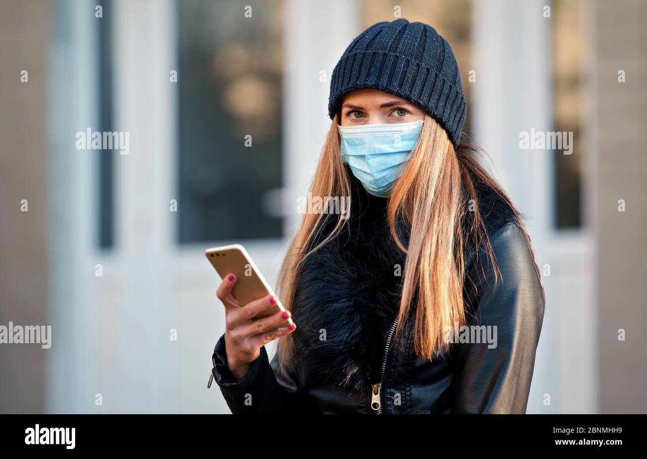 Junge Frau in schwarzer Jacke und warmer Mütze Hut, trägt Einweg-blauen  Virus Gesichtsmaske, Überprüfung ihres Handys. Coronavirus covid-19  Ausbruch vor Stockfotografie - Alamy