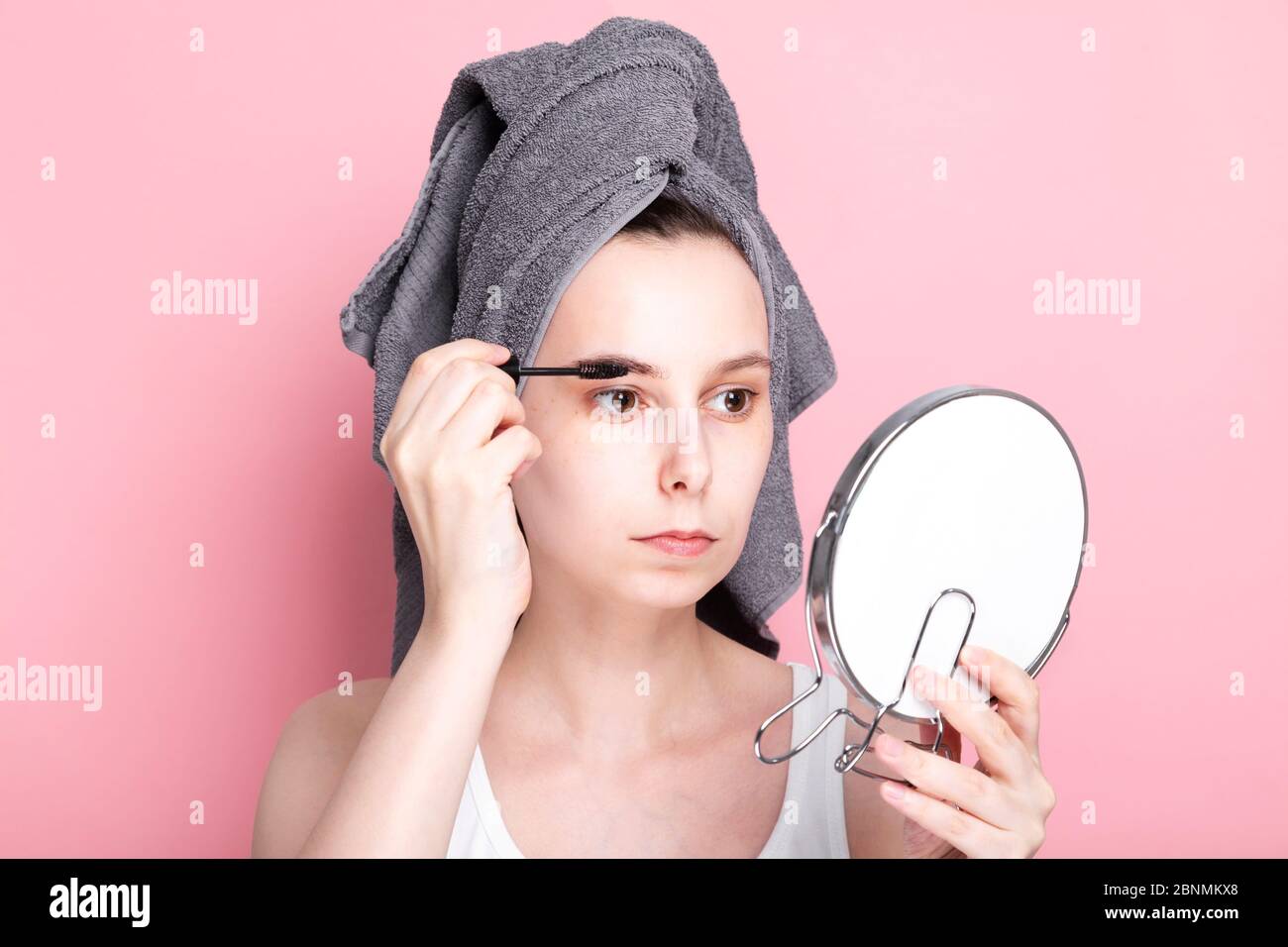 Junge Frau mit Handtuch auf dem Kopf malt ihre Augenbrauen auf sich selbst und sieht im Spiegel aus. Konzept Spa Tag zu Hause und in Quarantäne Stockfoto
