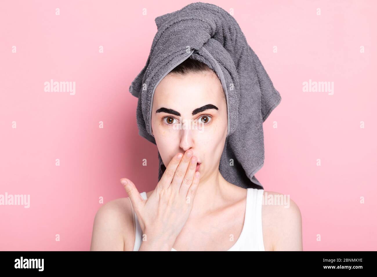 Junge Frau mit Handtuch auf dem Kopf malt ihre Augenbrauen auf sich. Von Überraschung bedeckt seinen Mund mit seiner Hand, etwas schief gelaufen. Konzept Spa Tag Stockfoto