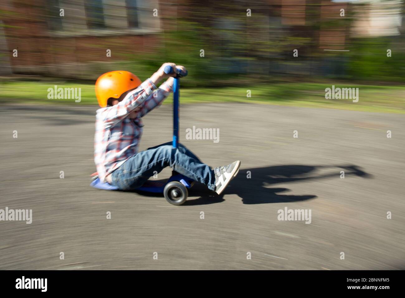 Junge draußen trägt orange Helm beschleunigt Auffahrt auf blau Roller Stockfoto