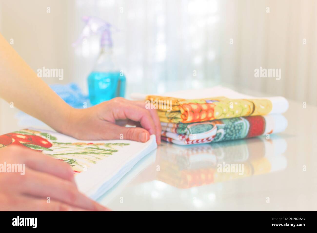 Hausarbeiten im Innenbereich. Hände einer Frau, die bunte Küchentücher auf einem weißen Tisch in der Nähe eines hellblauen Sprays faltet. Reinigungsprodukte. Hausarbeit. Reinigungsvorgang Stockfoto