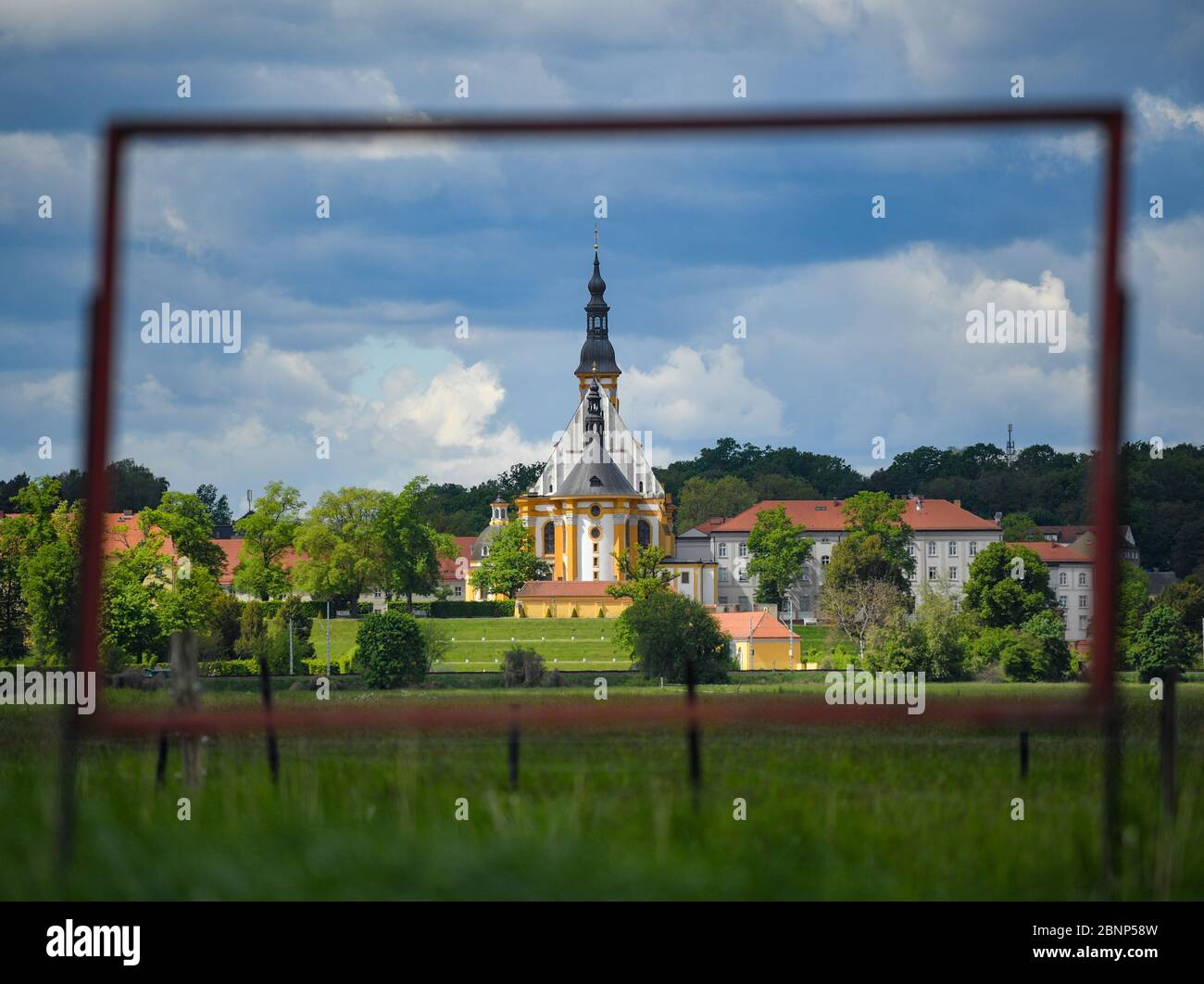 12. Mai 2020, Brandenburg, Neuzelle: Blick durch einen leeren Rahmen zur katholischen Kirche des Klosters Neuzelle. Im Klostergarten Neuzelle in der oder-Spree wird seit mehreren Monaten an der dritten und letzten Bauphase zur Sanierung der barocken Anlage gearbeitet. Zuvor waren in diesem Gebiet die Lauben eines Schottzartens aus DDR-Zeiten abgerissen worden. Bis 2022 wird hier eine Baumschule mit Gewächshaus, Nutzgarten und Kräutergarten sowie symmetrisch angeordnete Blumenbeete mit Hecken und Obstbäumen geschaffen. Seit 1998 wird an den Almos gearbeitet Stockfoto