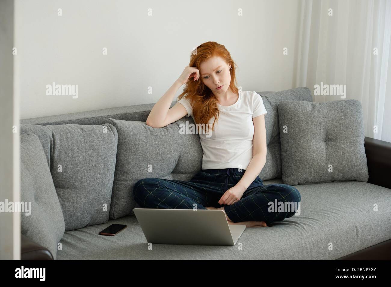 Ein rothaariges Mädchen in Heimkleidung sitzt mit gekreuzten Beinen auf der Couch und sieht sich vor ihr erzärtlich den Laptop an Stockfoto