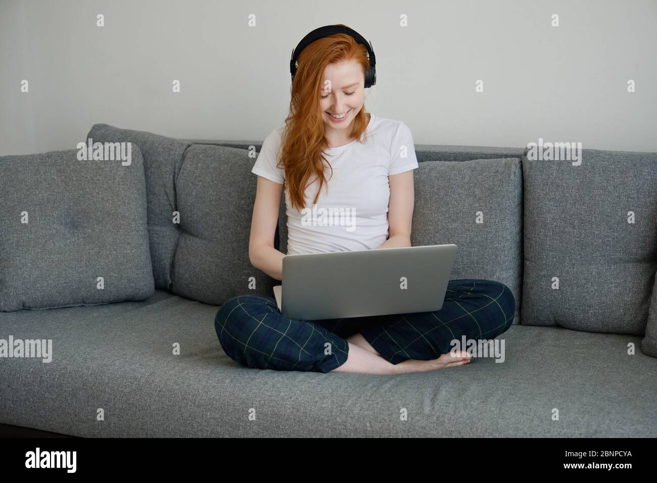 Rotschopf Mädchen sitzt auf einem Sofa in Heimkleidung mit einem Laptop auf gekreuzten Beinen und lächelnd, Blick nach unten Stockfoto