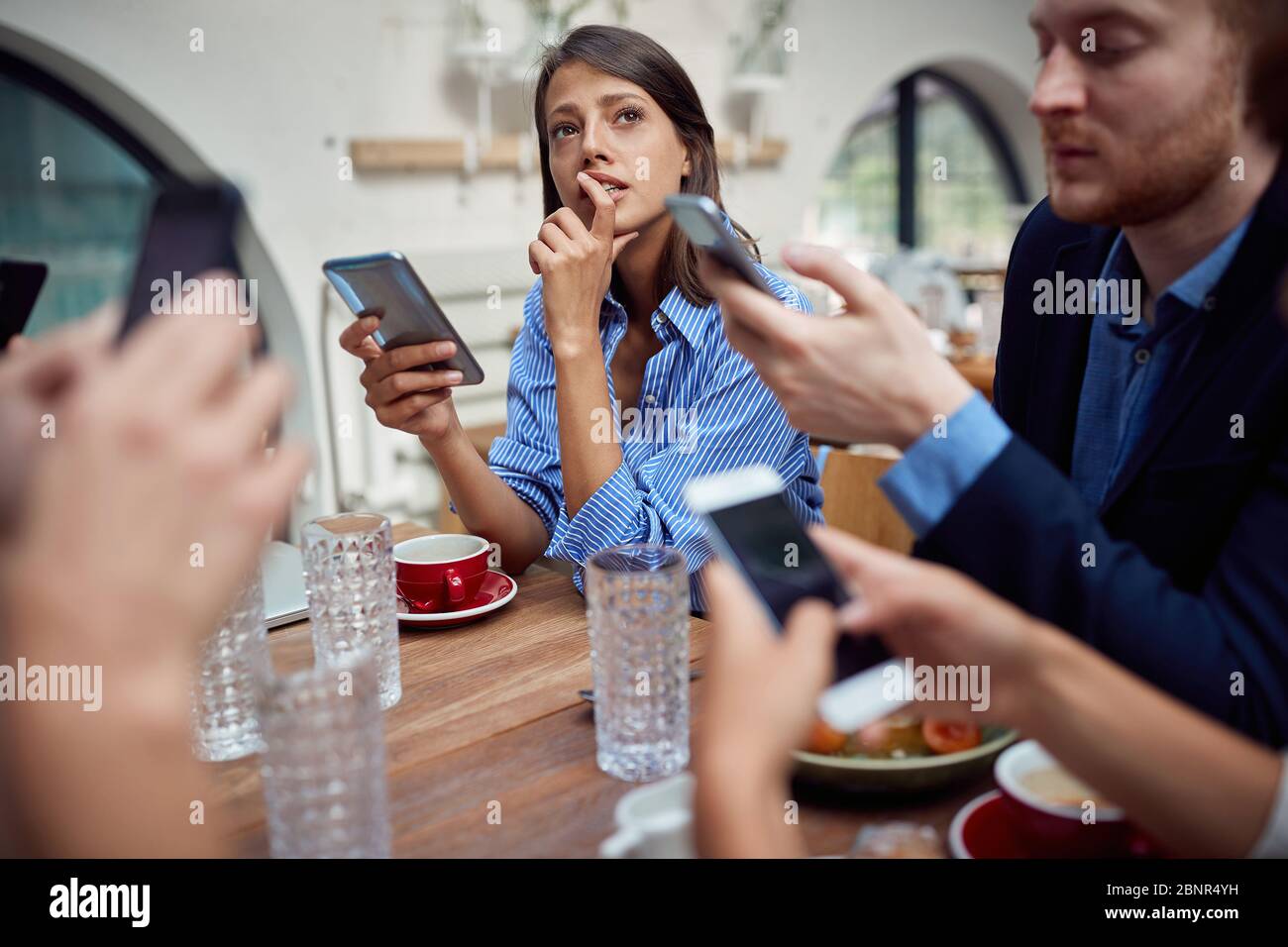 Junge kaukasische Frau fehlt in Gedanken auf unsoziale Mittagessen im Restaurant, wo jede Person mit dem Handy und nicht miteinander reden. Moderne te Stockfoto