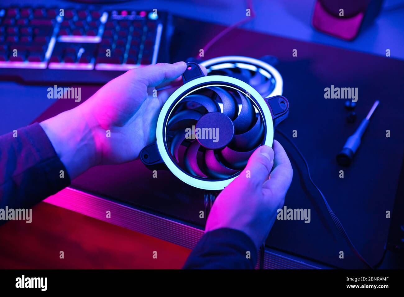 Die Hände des Mannes halten modernen Computerventilator mit blauem Licht mit Tastatur und Lautsprecher im Hintergrund. Stockfoto